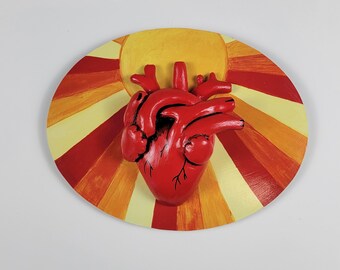 Könnte beinhalten: Eine dreidimensionale Skulptur eines roten anatomischen Herzens auf einer ovalen Platte. Die Platte zeigt ein gemaltes Sonnenstrahlenmuster in abwechselnden Gelb-, Orange- und Rottönen. Das Herz weist sichtbare Venen und Arterien auf.