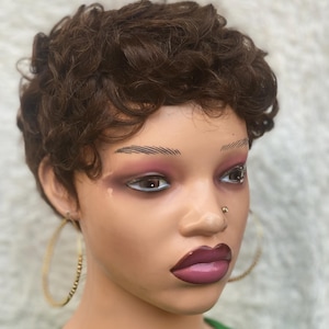 Peut inclure: Tête de mannequin avec une perruque courte et bouclée marron. Le mannequin porte un piercing au nez et de grandes créoles dorées. Les lèvres sont d'un rose foncé et les sourcils sont foncés. Le mannequin porte un haut vert.