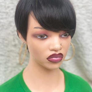 Peut inclure: Tête de mannequin avec une perruque courte noire avec une frange sur le côté. Le mannequin a un maquillage complet, y compris un rouge à lèvres rose foncé et des créoles dorées. Le mannequin porte un t-shirt vert à col en V.