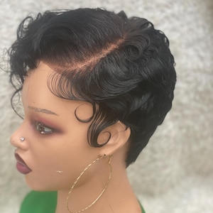 May include: A black, short-hair wig styled with finger waves, set on a mannequin head. The wig features a natural-looking hairline and is paired with large gold hoop earrings. The mannequin is wearing a green top.