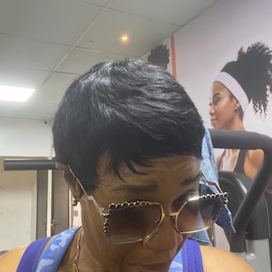 Puede incluir: Una mujer con gafas de sol cuadradas con montura dorada y adornos decorativos. Las lentes son tintadas y la mujer lleva una camiseta azul. El fondo sugiere un entorno de gimnasio.