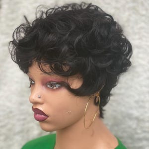 Peut inclure: Perruque courte et bouclée noire, coiffée avec une raie sur le côté. La perruque est sur une tête de mannequin avec un haut vert et des créoles dorées. Le mannequin a un piercing au nez et du rouge à lèvres foncé.