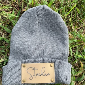 May include: Gray knitted beanie with a brown leather patch that says "Strider" in cursive.