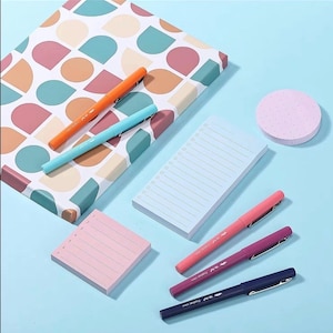 May include: A stationery set featuring a patterned notebook with geometric shapes in teal, pink, and beige, a rectangular checklist pad, a square notepad, a round dotted sticky note, and five colored fine-liner pens in orange, blue, pink, purple, and navy.