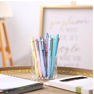 May include: A collection of retractable ballpoint pens in various pastel colors including yellow, pink, lavender, dark blue, light blue, and mint green. Each pen features a silver-toned metal clip and is stored upright in a clear glass tumbler.