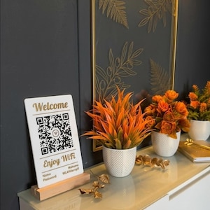 Könnte beinhalten: Eine dekorative Präsentation mit einem weißen Schild mit den Texten "Welcome" und "Enjoy WiFi", einem QR-Code und einem goldgerahmten Kunstwerk mit botanischen Designs. Orangefarbene Pflanzen in weißen Töpfen und ein goldenes Akzentstück vervollständigen das Arrangement.