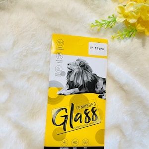 May include: A yellow and black package for a tempered glass screen protector for an iPhone 13 Pro. The package features a cartoon lion and the text "Tempered Glass Expertise New Experience".