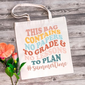 May include: Off-white canvas tote bag with the text "THIS BAG CONTAINS NO PAPERS NO GRADE & NO LESSONS TO PLAN #summertime" in various colors. The bag has a simple design with two handles and is placed on a wooden surface.