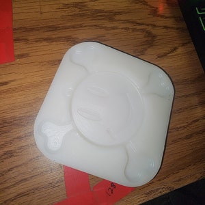 Puede incluir: Un molde blanco de forma cuadrada con un diseño de calavera y huesos cruzados. El molde tiene bordes redondeados y una hendidura circular en el centro, con una cara de calavera. El molde está sobre una superficie de madera.
