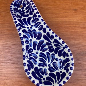 May include: A white ceramic spoon rest with a blue floral pattern. The spoon rest has a long, oval shape with a hole at the top for hanging. The blue design is a repeating pattern of stylized leaves and dots.