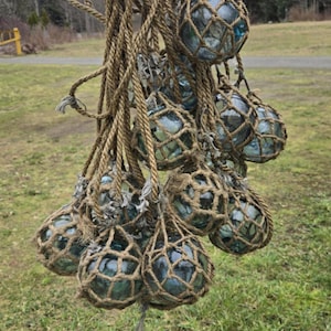 May include: A collection of blue glass fishing floats, each enclosed in a brown net, are hanging from thick brown rope. The floats are suspended in mid-air against a background of green grass.