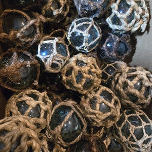 Può includere: Una collezione di galleggianti da pesca in vetro avvolti in spago naturale. I galleggianti sono di varie tonalità di blu e verde, e alcuni hanno un motivo a rete.