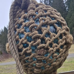 Może przedstawiać: Duża, okrągła, niebieska szklana boja osadzona w grubej, brązowej siatce z lin. Boja jest przymocowana do liny i wisi na drewnianym słupie.