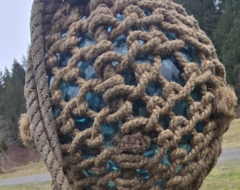 May include: A large, round, blue glass buoy encased in a thick, brown rope net. The buoy is attached to a rope and is hanging from a wooden post.