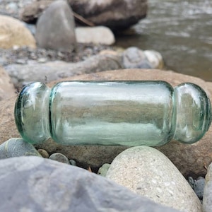 May include: A clear glass bottle with rounded ends, lying on a bed of gray rocks. The bottle has a greenish tint and is slightly weathered.