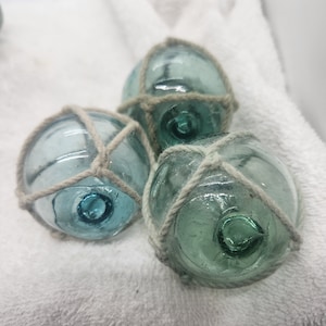 May include: Three glass fishing floats with a rope net design. The floats are a light blue-green colour and are sitting on a white fluffy surface.