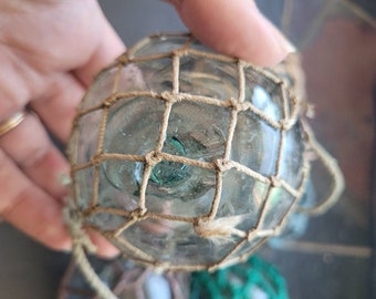 May include: A glass fishing float with a tan net covering the surface. The float is clear and has a green tint.