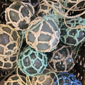 Peut inclure: Plusieurs boules de pêche en verre, de différentes nuances de bleu et de vert, enveloppées dans des filets tressés. Les boules sont suspendues par des cordes, créant une ambiance nautique. L'image présente une variété de tailles et de couleurs, évoquant un thème côtier.