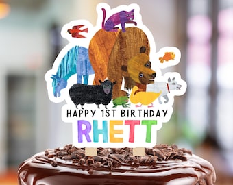 May include: A chocolate cake with a glossy chocolate glaze, topped with chocolate shavings. A colourful cake topper features a brown bear, a purple cat, and other animal illustrations. The text "HAPPY 1ST BIRTHDAY RHETT" is displayed on the topper.