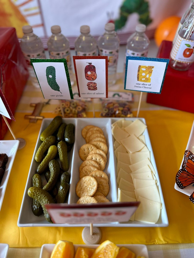 May include: A white serving tray with three compartments filled with snacks. The left compartment has green pickles, the middle compartment has round crackers, and the right compartment has thin slices of Swiss cheese. There are signs on sticks that say "one pickle", "one slice of salami", and "one slice of Swiss cheese".