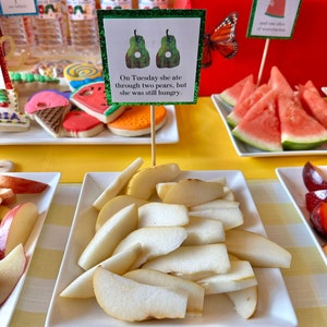 May include: A white plate with sliced pears on a yellow and white checkered tablecloth. A sign reads "On Tuesday she ate through two pears, but she was still hungry."