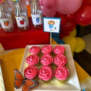 May include: A white platter with nine pink frosted cupcakes decorated with a swirl design. The cupcakes are arranged in three rows of three. A butterfly is on the platter. The platter is on a yellow surface. A sign with the text "one cupcake" is in the background.