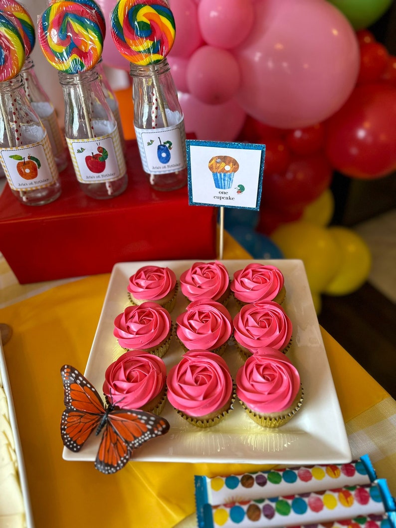 May include: A white platter with nine pink frosted cupcakes decorated with a swirl design. The cupcakes are arranged in three rows of three. A butterfly is on the platter. The platter is on a yellow surface. A sign with the text "one cupcake" is in the background.