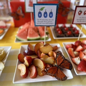 May include: A table setting for a children's party with a variety of fruits, including watermelon, strawberries, and plums. There are also butterfly decorations and signs with text from the book "The Very Hungry Caterpillar".