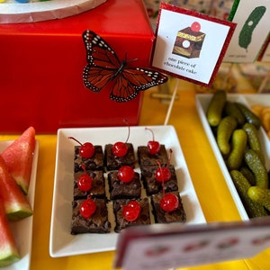 May include: A white plate with a selection of chocolate brownies topped with cherries. The brownies are arranged in a grid pattern. A sign reads "one piece of chocolate cake".