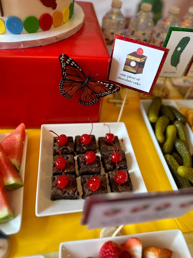 May include: A white plate with a selection of chocolate brownies topped with cherries. The brownies are arranged in a grid pattern. A sign reads "one piece of chocolate cake".
