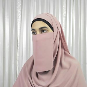 Może przedstawiać: Niqab w kolorze pudrowego różu z czarną lamówką wokół otworu na twarz. Niqab wykonany jest z miękkiej, przewiewnej tkaniny.