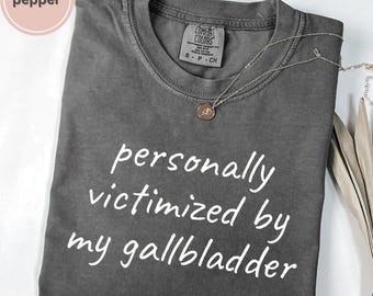 May include: A gray Comfort Colors t-shirt with the text "personally victimized by my gallbladder" in white. A silver necklace with a small pendant is draped across the shirt.