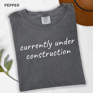 May include: A gray t-shirt with the text "currently under construction" in white script. The shirt is a Comfort Colors brand, and the tag is visible. The shirt is folded and appears to be on a white surface.