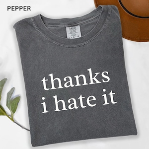 May include: Pepper-colored t-shirt with the text "thanks i hate it" in white. The shirt is a Comfort Colors brand and has a crew neck. The shirt is folded and has a relaxed fit.