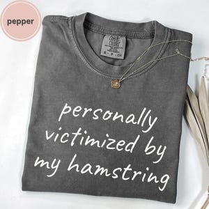 May include: A gray Comfort Colors t-shirt with the text "personally victimized by my hamstring" in white. A silver necklace with a small pendant is draped across the shirt. The shirt is folded on a white surface.