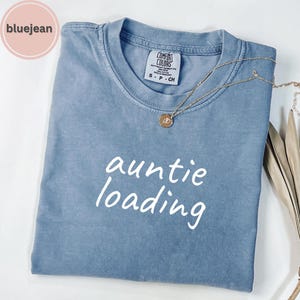 May include: A blue t-shirt with the words "auntie loading" in white script. The shirt has a crew neck and short sleeves. A gold necklace with a pendant rests on the shirt. The shirt is folded neatly.