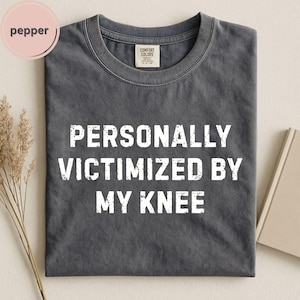 May include: A charcoal gray short-sleeve t-shirt featuring the white distressed text PERSONALLY VICTIMIZED BY MY KNEE printed on the front. The shirt has a crew neckline and a Comfort Colors brand label on the inside collar area.