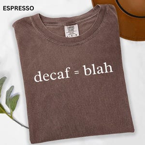 May include: Espresso-colored t-shirt with the text "decaf = blah" in white. The shirt is folded and has a Comfort Colors tag. A brown hat is partially visible in the background.