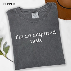 May include: A gray Comfort Colors t-shirt with the text "I'm an acquired taste" in white. The shirt is folded and has a crew neck. The word "PEPPER" is in the top left corner.