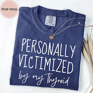 May include: A navy blue t-shirt with white text that reads PERSONALLY VICTIMIZED by my Thyroid. The shirt features a crew neckline and a Comfort Colors brand label at the inner collar. A thin gold-toned chain necklace with a small round pendant rests on top.
