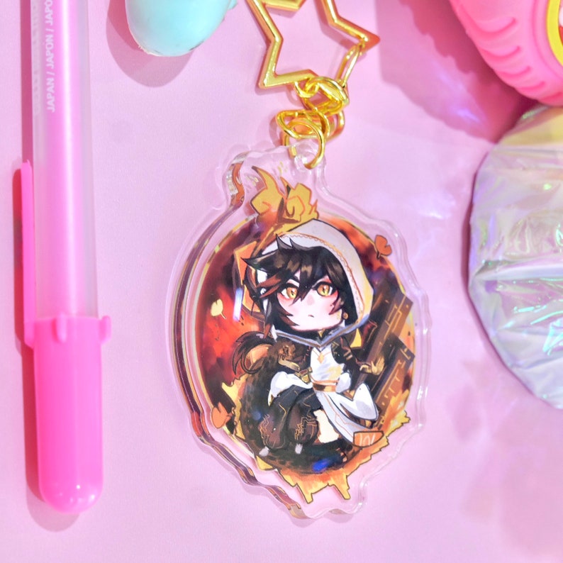 May include: A round, clear acrylic keychain featuring an anime-style character with black hair, a white hooded cloak, and yellow eyes. The character is standing in a fiery, orange and red background with a gold trim.