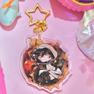 May include: A clear acrylic keychain with a gold star charm. The keychain features a cartoon illustration of a character with black hair, wearing a white hooded cloak and sitting on a golden background.