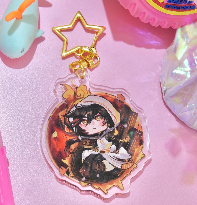 May include: A clear acrylic keychain with a gold star charm. The keychain features a cartoon illustration of a character with black hair, wearing a white hooded cloak and sitting on a golden background.