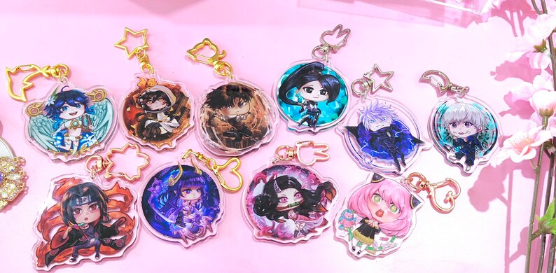 May include: A set of ten colourful anime character keychains. Each keychain features a different character with unique details and a clear background. The keychains are attached to gold-coloured chains with various charms.