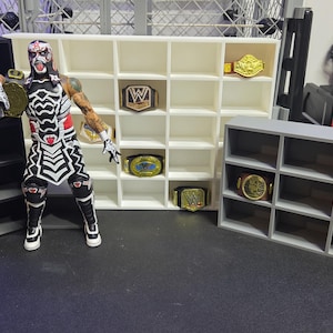 Könnte beinhalten: Ein Set aus schwarzen, weißen und grauen Kunststoff-Regalen zur Präsentation von Miniatur-Wrestling-Meisterschaftsgürteln. Das Set besteht aus einem hohen schwarzen Regal, einem breiten weißen Regal mit mehreren Fächern und einem kleineren grauen Regal.