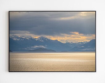 May include: A panoramic view of a mountain range with snow-capped peaks, seen from across a calm body of water. The sky is a mix of gray and white clouds, with a hint of golden light breaking through.