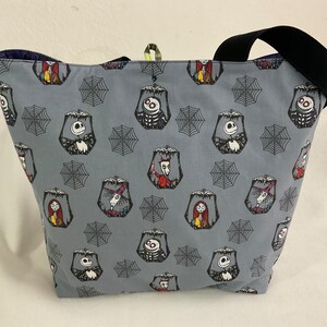 May include: A gray tote bag featuring a repeating pattern of characters and spiderwebs. The bag has a black strap and a dark blue interior. The characters include Jack Skellington, Sally, and other figures from the movie.