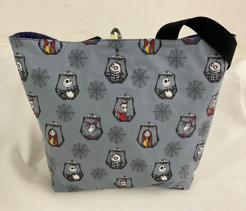 May include: A gray tote bag featuring a repeating pattern of characters and spiderwebs. The bag has a black strap and a dark blue interior. The characters include Jack Skellington, Sally, and other figures from the movie.