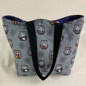 May include: Gray tote bag featuring a repeating pattern of characters and spiderwebs. The bag has black handles and a dark purple interior. The characters include Jack Skellington, Sally, and other figures from the movie. The bag is made of fabric.