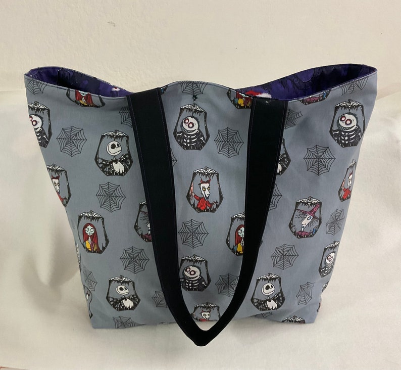 May include: Gray tote bag featuring a repeating pattern of characters and spiderwebs. The bag has black handles and a dark purple interior. The characters include Jack Skellington, Sally, and other figures from the movie. The bag is made of fabric.